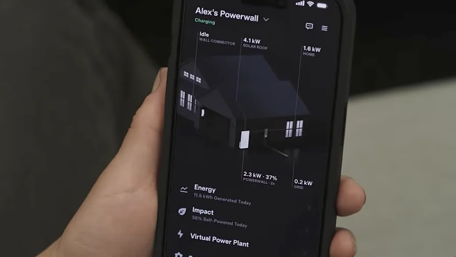 Hand holding a smartphone displaying a home energy app with a house graphic and real-time power flow data, including solar generation, battery storage, home usage, and grid connection.