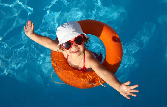 Kid swimming in a pool and wearing a floatie, sunglasses and a white hat.