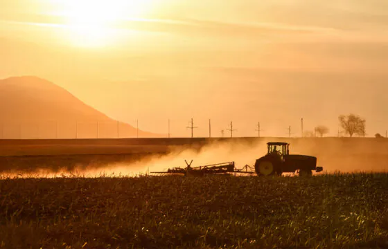 Tractor farming a field with a sunset, mountain, and power lines in the background.